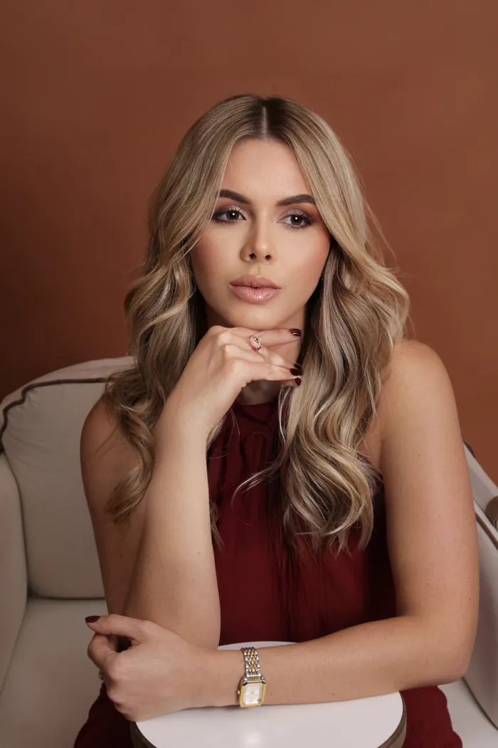 Dra. Cátia Jaine de vestido bordô, sentada, com a mão no rosto